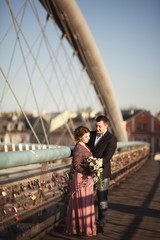 Fototapeta premium Stylish loving wedding couple, groom, bride with pink dress kissing and hugging on a bridge at sunset