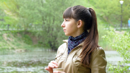 Beautiful young girl in the park drinking cup of takeaway coffee  from disposable cup. Portrait of attractive woman thoughtfully looks out over river and holding cup of tea  to warm up