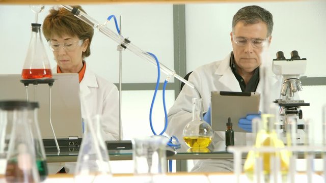 A distillation kit frames a female chemist conferring with her male counterpart working with a microscope and electronic tablet. Slow dolly movement past lab glassware in the foreground.