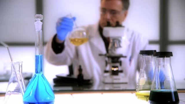 Focus remains on a volumetric flask and other lab glassware while a man working with a microscope works in the background.