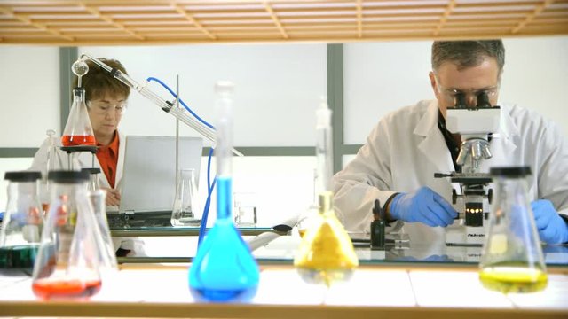 Laboratory glassware in the foreground give depth to the footage of two scientists working in their lab.