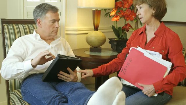A man enjoying what he is engaged in on his electronic tablet is interrupted by his wife who changes his mood by handing him cleaning materials.