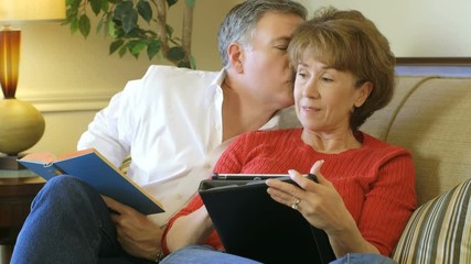 A mature couple sitting on a couch together one reading a book and the other using an electronic tablet pc. - Powered by Adobe