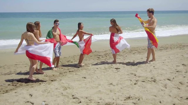 Group of Young Adult Friends Wearing Bathing Suits and Waving Flags from Various Countries While Standing Together on Sunny Beach with View of Ocean in Background.