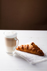 Glass of latte macchiato and French croissant on baking paper. Vertical scene on white wooden background.