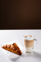 Glass of latte macchiato and French croissant on baking paper. Vertical scene on white wooden background.