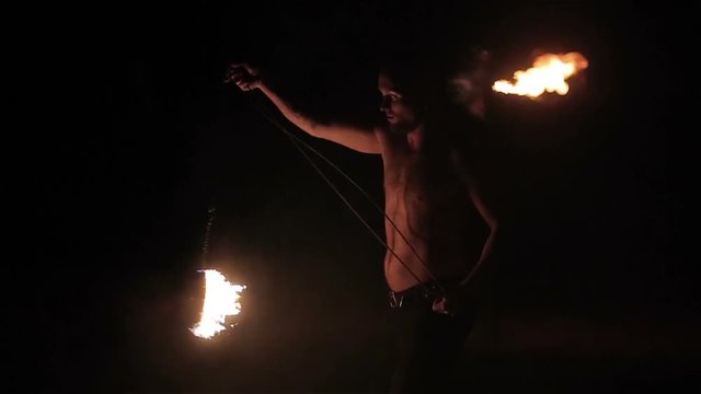 Fire show performance. Male fire performer spinning burning fire rope dart poi on long rope around himself. Slow motion