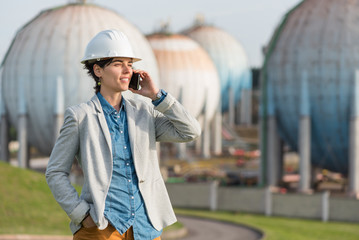 successful independent engineer smiling woman talking on the phone on industrial area with safety helmet. Pioneer woman at work with spherical tanks.