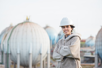 successful independent engineer smiling woman on industrial area with safety helmet crossing arms. Pioneer woman at work with spherical tanks.