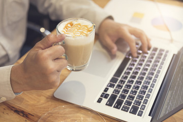 man's hand holding cup of hot latte coffee working with laptop c