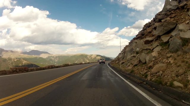Cliff on one side and boulder strewn mountainside on the other as a Rocky Mountain National Park road unfolds in front of the camera. Car mounted camera footage.