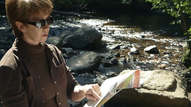 Scene changes focus from a stream in the forest to a mature woman using a paper map to determine her whereabouts.