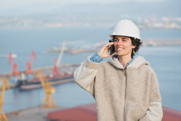 successful independent engineer smiling woman on industrial harbor with safety helmet talking on the phone. Pioneer woman at work.