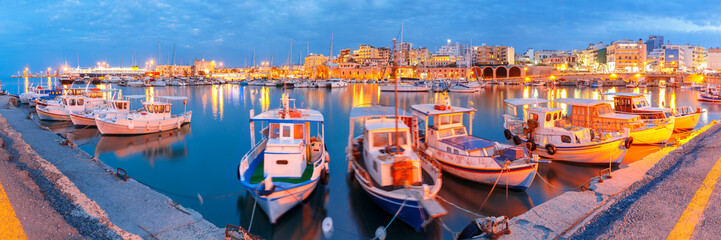 Panorama of Old harbour with fishing boats and marina during twilight blue hour, Heraklion, Crete, Greece. Boats blurred motion on the foreground.