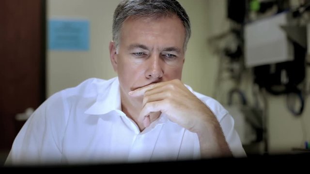 An IT admin in his office looking at information on his computer monitor with cables and wires hanging on the wall behind him.