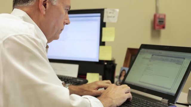 A businessman in a rather industrial looking office uses his laptop verses his desktop computer to type an email.