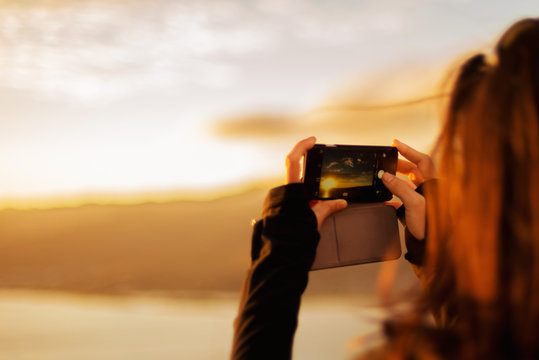 Brunette woman taking photo of sunset with smart phone, with focus on the screen