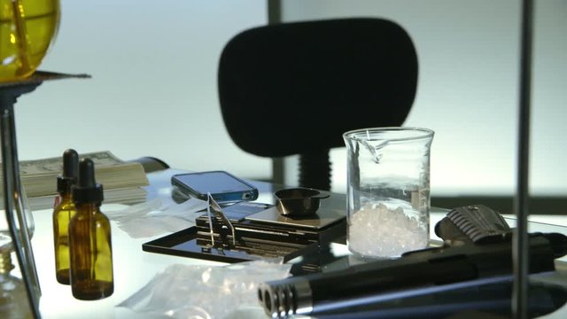 Panning down from substance boiling in laboratory flask to a gun and other paraphernalia of sinister looking scene.