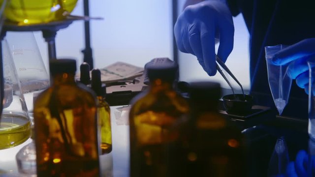 Scene moves across sinister looking laboratory paraphernalia where a man with gun nearby is working with a crystallized substance.