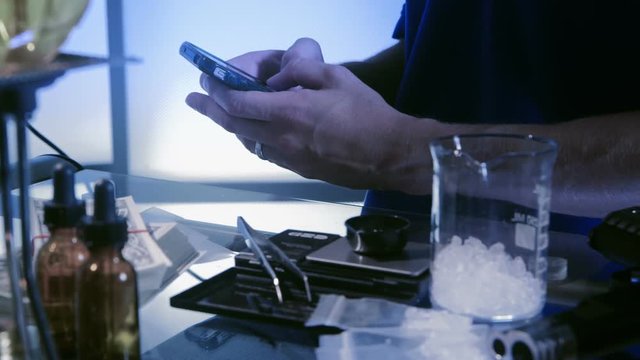 A man sitting at a table with laboratory paraphernalia used to make illegal drugs texting on his smart phone.