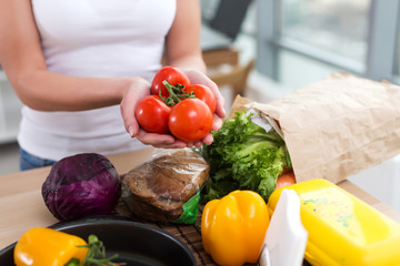 Female hands of a caucasian cook holding red tomato bunch over kitchen worktop with fresh grocery and rye bread on it. 