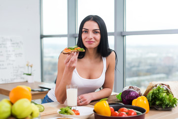Close-up portrait of smiling woman eating diet vegetarian sandwich with vegetables for breakfast in morning, looking at camera. 