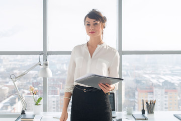 Attractive ambitious businesswoman standing in modern office, holding paper folder, looking at...