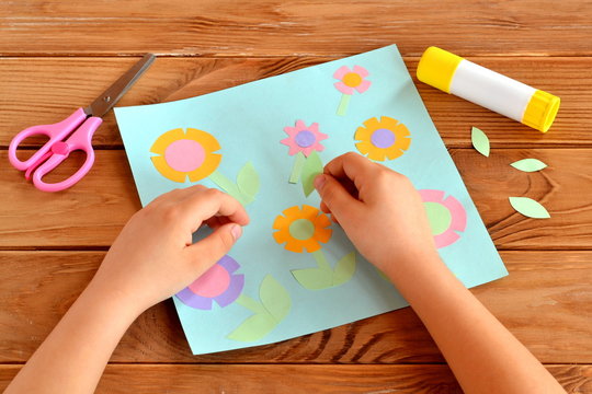 Child doing a card with flowers. Child holds a paper leaf in his hands and sticks it. Glue, scissors, greeting card on a wooden table. Paper flower craft for kids. Children's art project.