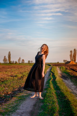 Girl in black dress is dancing on the field road barefoot. happy woman outdoors. Sunset. meadow.