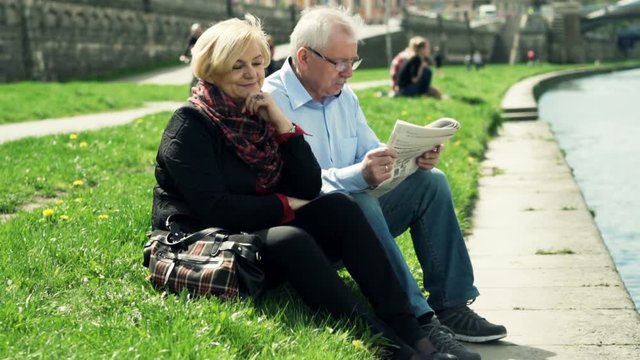 Mature couple relaxing and reading newspaper sitting on grass near city river
