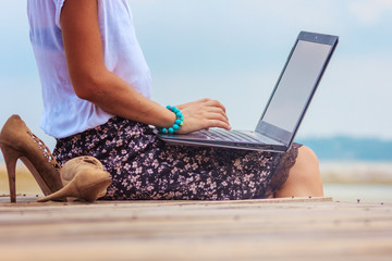 Young woman sitting with a laptop on the wooden platform edge at the sea. Lady freelancer in office...