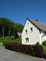 Wohnhaus mit spitzem Giebel und Hecke an einem Berghang vor blauem Himmel im Sonnenschein im...