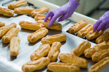 Eclair shells beside conveyor belt. Hand in glove touching eclairs. Worker selects best eclairs. Dough products made at factory.