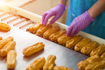 Hands in gloves touching eclairs. Eclair shells on narrow conveyor. Employee of confectionery plant. Worker responsible for quality.