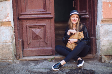 Portrait of a stylish and beautiful teen girl in the hat is posing and laughing and holding a red cat in her arms