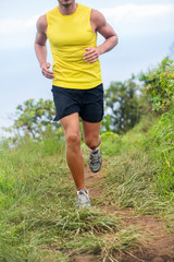 Athletic legs of fit runner man training cardio workout on trail mud grass path in summer nature outains. Sports man lower body crop for thighs and knees concept wearing shorts and running shoes.