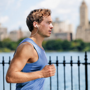 Urban young man running in city park listening to music with wireless bluetooth in-ear earphones living a healthy active lifestyle. Male runner blue top working out cardio exercise workout in summer.