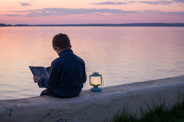Fototapeta premium child reading a book with lantern at sunset near water