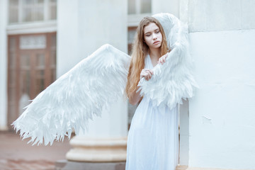 Young woman with white wings is standing near the column. The girl hid wing