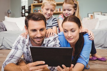 Fototapeta premium Parents holding digital tablet lying on floor while kids sitting on their back at home