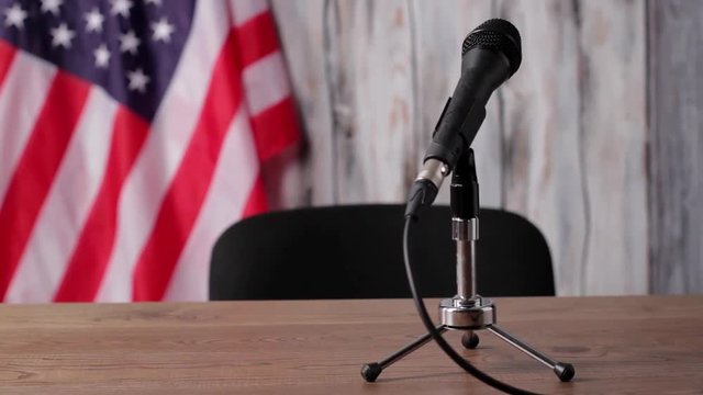 American flag, table and microphone. Banner behind desk with microphone. Sound equipment is ready. News broadcast will soon begin.