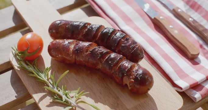 Two char grilled sausages with fresh rosemary and a tomato on a wooden picnic table outdoors  close up view