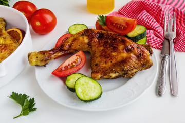 Roasted chicken legs cooked with sauce from orange, honey and olive oil. On white plate with fresh vegetables. White table.