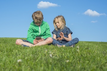 Naklejka na ściany i meble Happy blond children using smartphone (watching movie or playing game) sitting on the grass. Brother and sister. Summertime