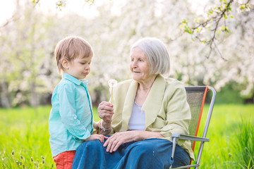 Little boy blowing dandelion seeds while his great grandmother is holding a flower
