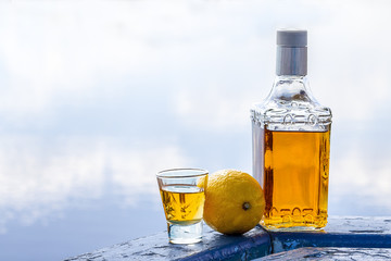 Bottle tequila and tumbler with lemon on shore of lake