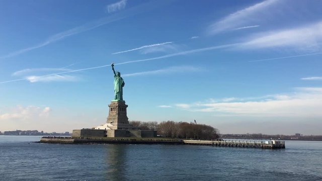 Liberty Island shown beneath beautiful bold blue sky over tranquil reflective Hudson bay waters.

