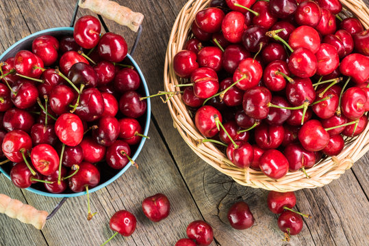Red cherries in metal bucket and basket on a wooden background.
