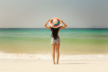 Beautiful woman in sun hat and bikini standing with arms raised to her head enjoying looking view...