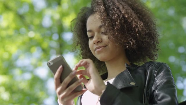 Beautiful young girl with dark curly hair using her cell phone, outdoor. Portrait of relaxed young lady in a summer park reading a text message on her mobile phone.